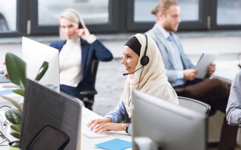 selective focus of smiling arabic call center operator in hijab in headset working on computer at